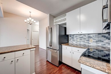 This modern kitchen features sleek white cabinets, granite countertops, and a stylish tiled backsplash. It includes stainless steel appliances and dark hardwood floors, with a bright, open layout enhanced by a contemporary chandelier.