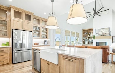 Kitchen with light brown cabinetry, appliances with stainless steel finishes, light stone counters, high vaulted ceiling, and pendant lighting