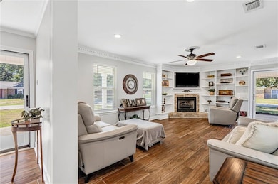 Living room featuring dark wood-style flooring, crown molding, a fireplace with raised hearth, recessed lighting, and a ceiling fan