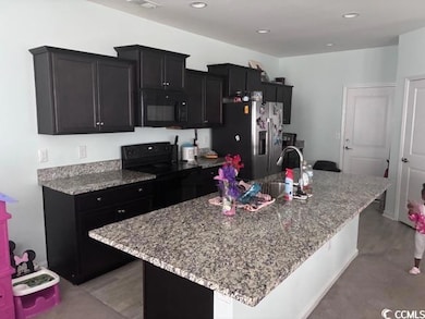 Kitchen with dark cabinets, black appliances, a center island with sink, light stone countertops, and recessed lighting