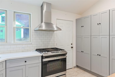 Kitchen with gray cabinets, stainless steel gas stove, wall chimney range hood, decorative backsplash, and light stone counters