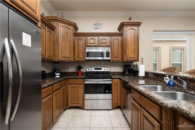 Kitchen with appliances with stainless steel finishes, crown molding, brown cabinetry, light tile patterned floors, and dark stone counters