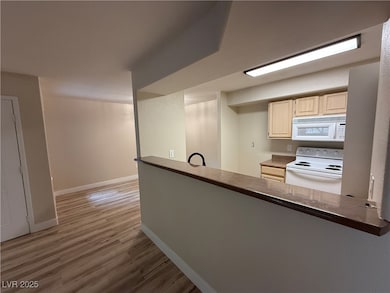 Kitchen with light brown cabinetry, dark countertops, white appliances, baseboards, and wood finished floors