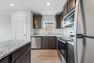 Kitchen featuring appliances with stainless steel finishes, light wood-style floors, dark brown cabinetry, light stone countertops, and recessed lighting