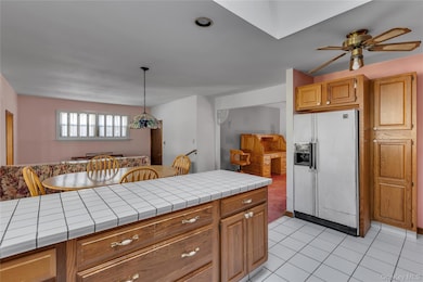 Kitchen featuring tile counters, white refrigerator with ice dispenser, brown cabinetry, light tile patterned flooring, and decorative light fixtures