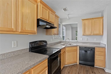 Kitchen featuring black appliances, light brown cabinetry, under cabinet range hood, and wood finished floors