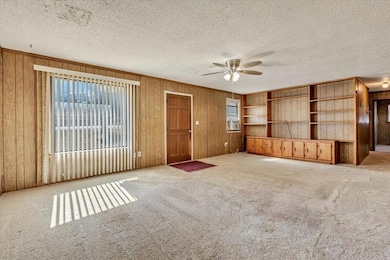 Unfurnished living room featuring light carpet, a ceiling fan, a textured ceiling, wooden walls, and crown molding