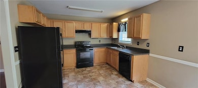 Kitchen featuring black appliances, under cabinet range hood, light brown cabinetry, dark countertops, and stone finish floors