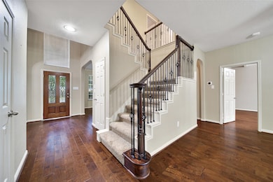 This inviting entryway features warm hardwood floors, a beautifully crafted staircase with wrought iron balusters, and a wooden front door with decorative glass. The open layout includes arched doorways leading to adjacent rooms, creating a welcoming and elegant atmosphere.