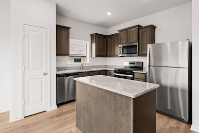 Kitchen with stainless steel appliances, a center island, light stone countertops, dark brown cabinetry, and light wood finished floors