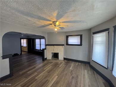 Unfurnished living room featuring a ceiling fan, dark wood-type flooring, a brick fireplace, a textured ceiling, and healthy amount of natural light