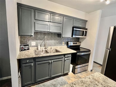 Kitchen featuring gray cabinetry, stainless steel appliances, tasteful backsplash, a textured wall, and light stone countertops