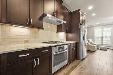 Kitchen featuring dark brown cabinets, light stone countertops, under cabinet range hood, appliances with stainless steel finishes, and tasteful backsplash
