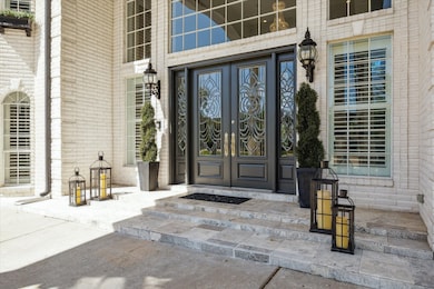 This elegant entrance features a grand double door with intricate glass designs, framed by tall windows. The area is accented with stylish lanterns, set against a light brick exterior.