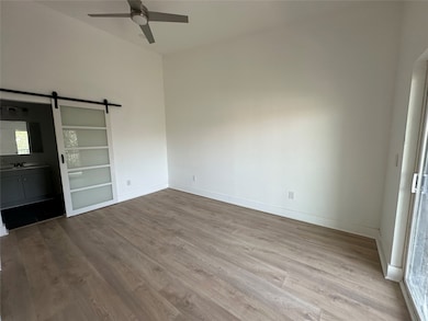 Spacious room with light wood flooring, a modern ceiling fan, and a sliding barn door leading to a bathroom. Ample natural light enters through large windows.