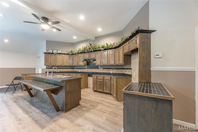Kitchen featuring dark countertops, light wood-type flooring, a ceiling fan, an island with sink, and a kitchen bar