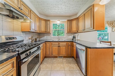 Kitchen with stainless steel appliances, under cabinet range hood, brown cabinetry, a peninsula, and backsplash