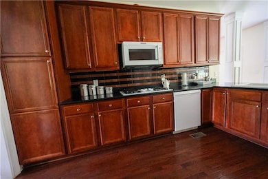 Kitchen with white appliances, dark wood finished floors, dark countertops, tasteful backsplash, and brown cabinets