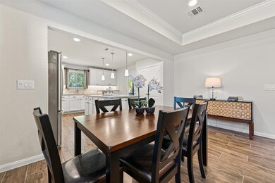 Contemporary dining area with a wooden table and black chairs, leading into a bright kitchen with white cabinetry and modern lighting. The space features wood-style flooring and a sideboard with a lamp for added warmth. Large dining area can accommodate even more seating for those large gatherings. (Furniture available for purchase)