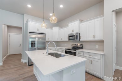 Kitchen featuring stainless steel appliances, white cabinetry, light wood-style floors, hanging light fixtures, and an island with sink