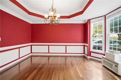 Empty room with a raised ceiling, a chandelier, ornamental molding, light wood-style flooring, and wainscoting