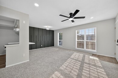 Bright, open living space featuring carpeted flooring, a modern black ceiling fan, and a large window with blinds for natural light. The room connects to a kitchen area with a partial wall, creating a cozy and functional layout.