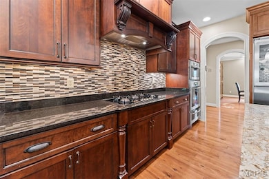 Kitchen featuring dark stone counters, arched walkways, light wood-style flooring, stainless steel appliances, and premium range hood