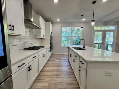 Kitchen with white cabinets, wall chimney range hood, a kitchen island with sink, light stone countertops, and decorative light fixtures