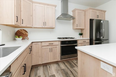 Kitchen featuring light countertops, stainless steel appliances, light brown cabinetry, light wood finished floors, and wall chimney range hood