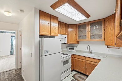 Kitchen with white appliances, brown cabinetry, light countertops, glass insert cabinets, and light carpet