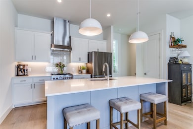 Kitchen with light wood-type flooring, a center island with sink, wall chimney exhaust hood, and stainless steel fridge