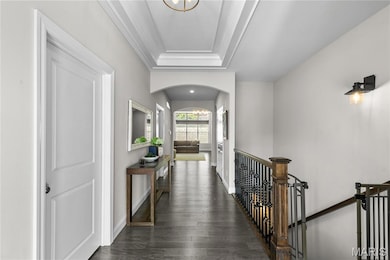 Corridor with a tray ceiling, dark wood finished floors, arched walkways, ornamental molding, and an upstairs landing