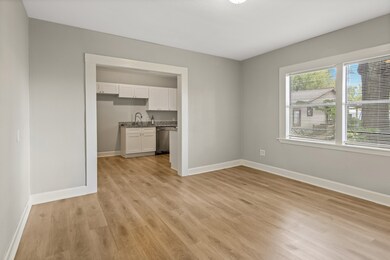 Living room showcases a bright, open room with light wood flooring and neutral walls. It features a large window allowing natural light and an open entryway leading into a kitchen.