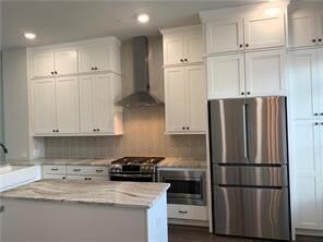 Kitchen with wall chimney range hood, freestanding refrigerator, white cabinetry, light stone counters, and black range