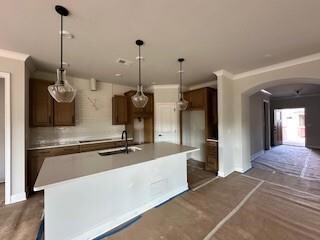 Kitchen featuring arched walkways, crown molding, a center island with sink, decorative light fixtures, and light countertops