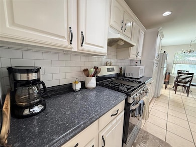 Kitchen featuring white appliances, under cabinet range hood, light tile patterned floors, tasteful backsplash, and white cabinets