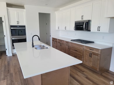 Kitchen featuring white cabinetry, double wall oven, dark wood-style flooring, a kitchen island with sink, and brown cabinetry