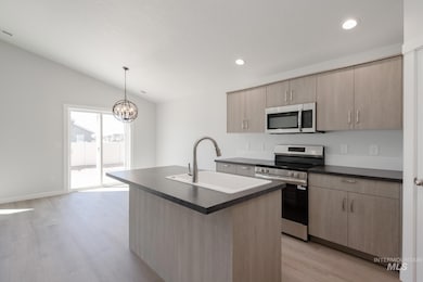 Kitchen featuring dark countertops, stainless steel appliances, an island with sink, light brown cabinetry, and vaulted ceiling