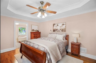 Bedroom with a tray ceiling, ornamental molding, wood finished floors, connected bathroom, and ceiling fan