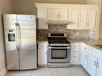 Kitchen with appliances with stainless steel finishes, under cabinet range hood, light stone countertops, backsplash, and white cabinets