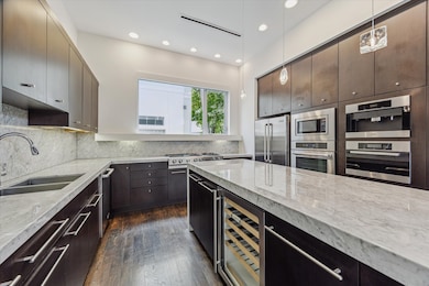 This modern kitchen features sleek dark custom cabinetry, marble countertops, and stainless steel appliances, including a built-in oven, microwave, and wine cooler. It offers ample storage and counter space, with a large window providing natural light.