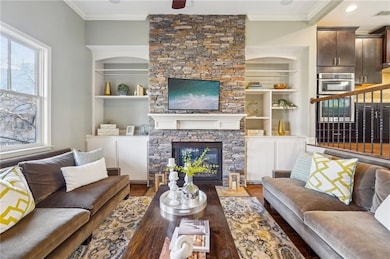 Living room with ornamental molding, a stone fireplace, wood finished floors, and built in features