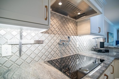 Kitchen with custom exhaust hood, black electric stovetop, decorative backsplash, white cabinets, and dark stone countertops
