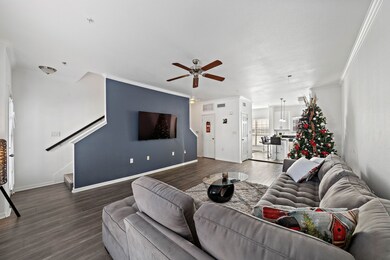 This spacious living area features an open concept design with a modern kitchen, cozy sectional, and a wall-mounted TV. The room is bright with a ceiling fan and stylish flooring, and a staircase leads to the upper level. A decorative Christmas tree adds a festive touch.