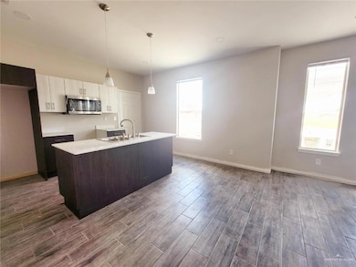 Kitchen featuring light countertops, stainless steel microwave, decorative light fixtures, dark wood-type flooring, and a center island with sink