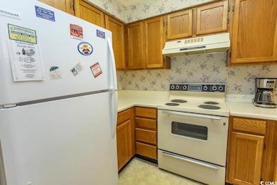 Kitchen with white appliances, under cabinet range hood, wallpapered walls, light countertops, and brown cabinetry