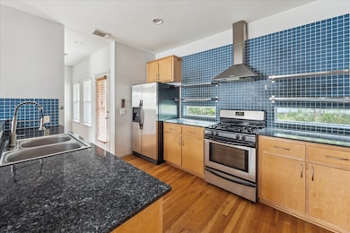 The modern kitchen boasts sleek black granite countertops, stainless steel appliances, natural wood cabinets, a gas stove, and a stylish blue tile backsplash. It's bright and airy with plenty of natural light.