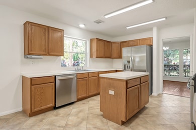 This bright, spacious kitchen features modern stainless steel appliances, ample wooden cabinetry, and a central island. Natural light floods through a large window, highlighting the clean, tiled floor. Adjacent is a dining area with a view of the outdoors.