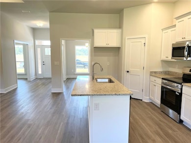 Kitchen featuring stainless steel appliances, light stone counters, white cabinetry, an island with sink, and dark wood finished floors