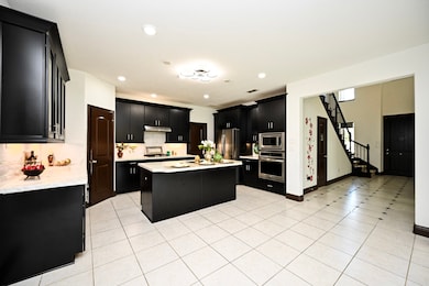 Spacious modern kitchen with sleek dark cabinetry and light countertops. Features  huge  island, stainless steel appliances, and an open layout leading to a tiled hallway with a staircase.